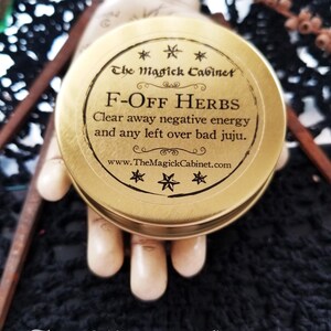 May include: A gold metal container with a black label that reads "The Magick Cabinet" and "F-OFF HERBS Clear away negative energy and any left over bad juju. www.TheMagickCabinet.com".