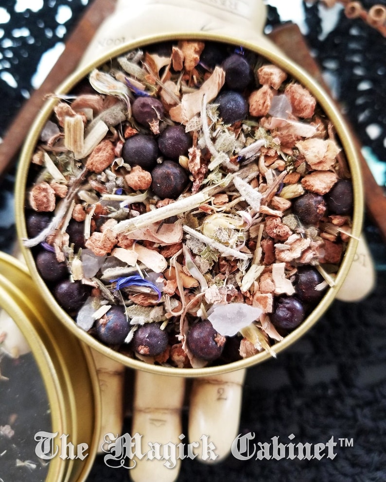 May include: A close-up of a round tin filled with a blend of dried herbs, including juniper berries, sage, and other botanicals. The tin is gold-colored and has a label that reads "The Magick Cabinet".