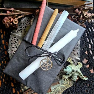 May include: A black package tied with black string, containing a white candle, incense sticks, and a selenite wand. A gold-colored pentagram charm hangs from the string. The package is on a decorative metal tray.