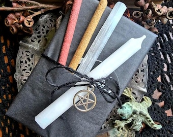 May include: A black package tied with black string, containing a white candle, incense sticks, and a selenite wand. A gold-colored pentagram charm hangs from the string. The package is on a decorative metal tray.