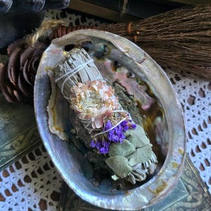 May include: An iridescent abalone shell holding a sage smudge stick wrapped in white string, adorned with a dried light pink flower, purple statice, and green eucalyptus leaves. The text "The Magick Cabinet" is printed on the surface below.