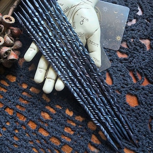 May include: A set of ten black twisted metal wands, approximately 10 inches long, are displayed on a black crocheted surface. The wands are arranged in a fan shape, with the tips pointing towards the right side of the image. The wands are made of a dark metal, and they have a twisted, or spiral, design. The wands are all the same length, and they are all the same width. The wands are arranged in a way that suggests they are for use in a magical or ceremonial context.