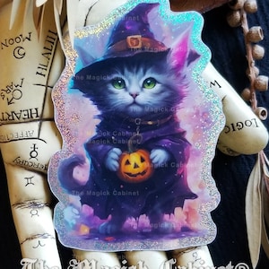 May include: A black cat wearing a witch's hat and holding a jack-o'-lantern. The cat is standing in front of a purple and pink background with stars. The image has a glittery, holographic effect.