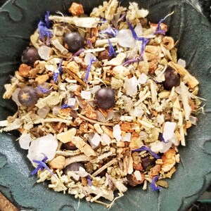 May include: A close-up of a black bowl filled with a mixture of dried herbs, flowers, and berries. The mixture includes juniper berries, dried lavender flowers, and other herbs. The bowl is on a dark surface.