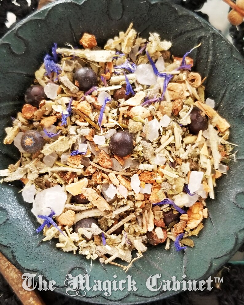 May include: A close-up of a black bowl filled with a mixture of dried herbs, flowers, and berries. The mixture includes juniper berries, dried lavender flowers, and other herbs. The bowl is on a dark surface.