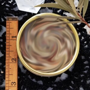 Closed tin of herbs showing the different ingredients. Next to a ruler showing the tin is 2.5 inches in diameter. Black lace tablecloth is in the background. The Magick Cabinet is watermarked at the bottom of the photo.