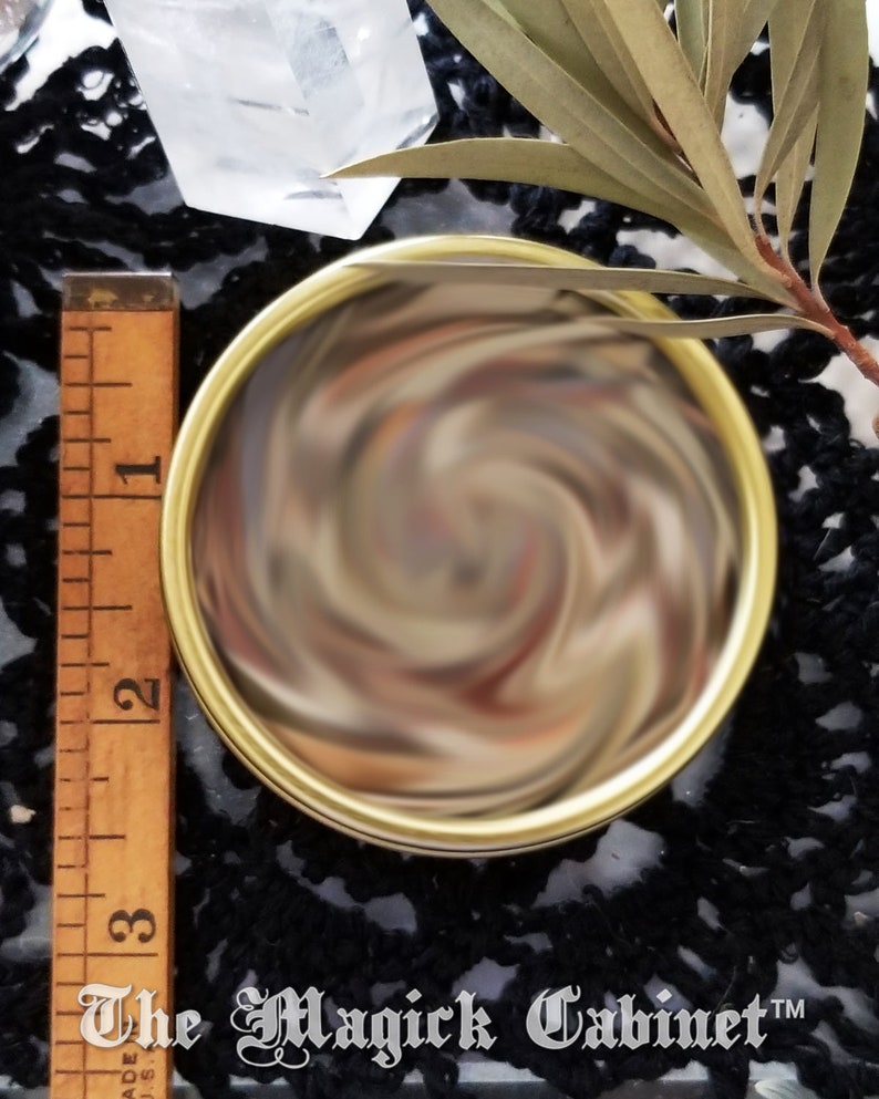Closed tin of herbs showing the different ingredients. Next to a ruler showing the tin is 2.5 inches in diameter. Black lace tablecloth is in the background. The Magick Cabinet is watermarked at the bottom of the photo.