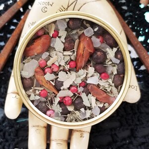 May include: A round metal tin filled with a mixture of dried herbs, spices, and berries. The tin is sitting on a hand with the words "The Magick Cabinet" written on the bottom.