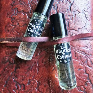 May include: Two small glass bottles with black caps and labels that say "The Magick Cabinet" and "Magically Infused Perfume". The bottles are sitting on a brown leather surface.