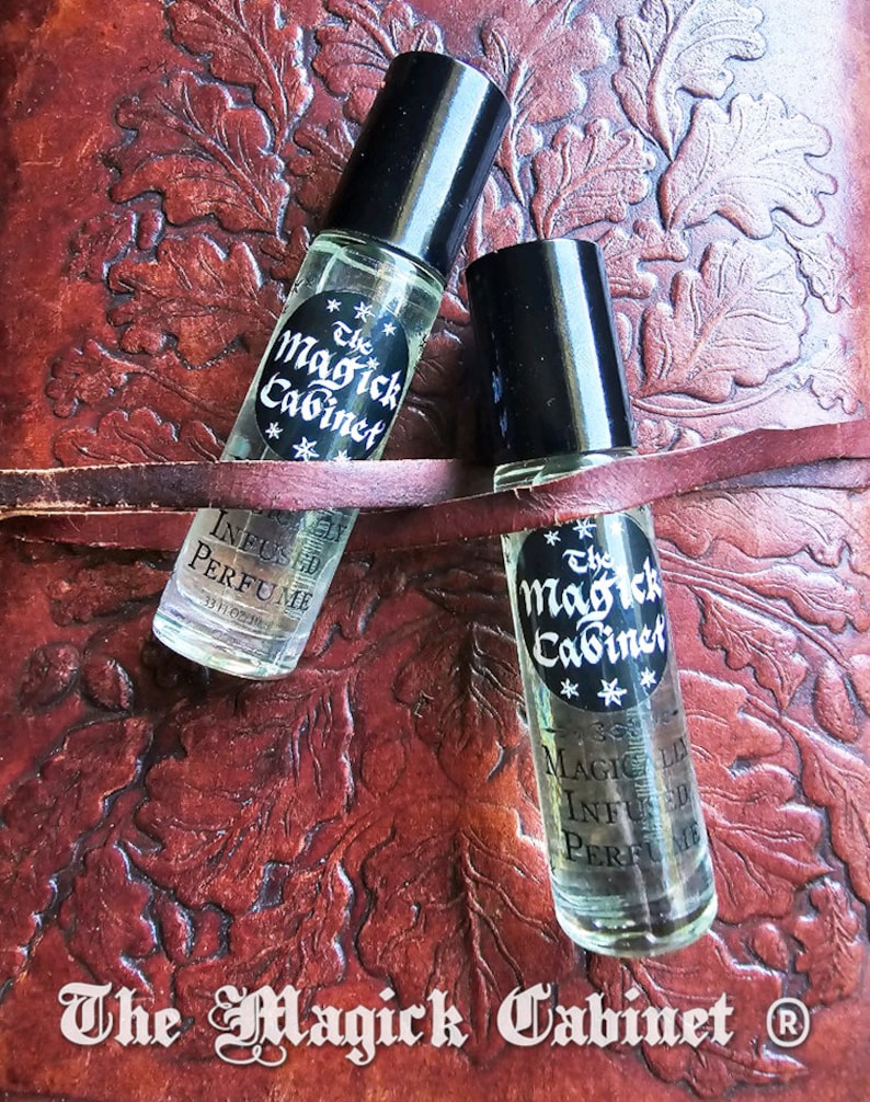May include: Two small glass bottles with black caps and labels that say "The Magick Cabinet" and "Magically Infused Perfume". The bottles are sitting on a brown leather surface.