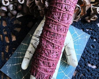 May include: A purple sage smudge stick wrapped in twine, resting on a white hand with a spiderweb design. The hand is on a blue and white card with the text "The Magick Cabinet" and a registered trademark symbol.