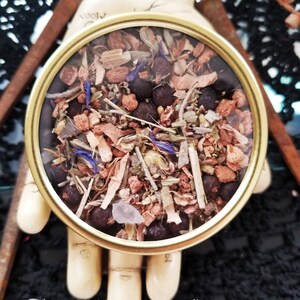 May include: A round, clear glass jar with a gold lid filled with a mixture of dried herbs, flowers, and berries. The jar is sitting on a wooden hand with the text "The Magick Cabinet&trade;" below it.