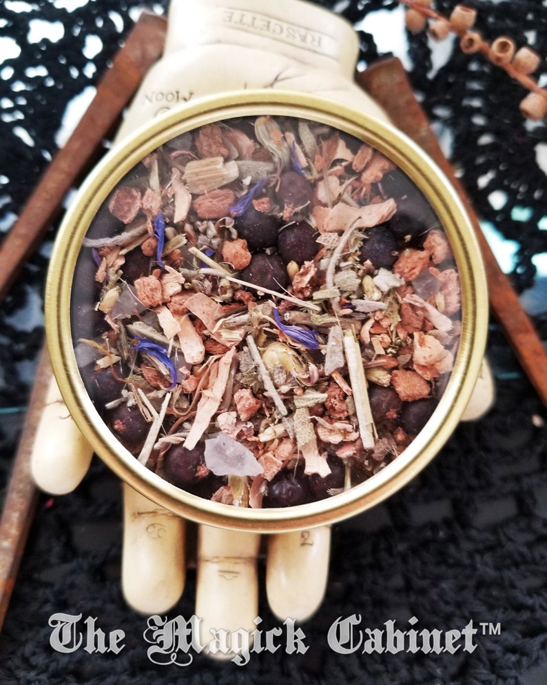 May include: A round, clear glass jar with a gold lid filled with a mixture of dried herbs, flowers, and berries. The jar is sitting on a wooden hand with the text "The Magick Cabinet&trade;" below it.