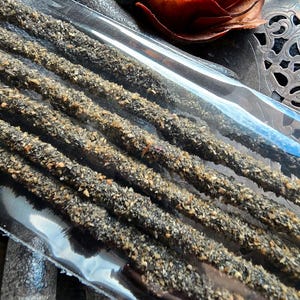 May include: Close-up of several incense sticks in a clear plastic package. The incense sticks are dark brown and covered in small, light-colored granules. A dried, reddish-brown flower and decorative metal piece are in the background. The text "The Magick Cabinet" is visible.