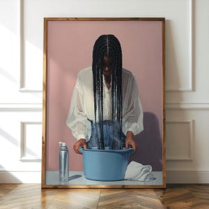 Puede incluir: Una impresión artística enmarcada representa a una persona con largas trenzas oscuras sobre una palangana azul. La persona lleva una blusa blanca y vaqueros. Un pulverizador y una toalla blanca están sobre la mesa. El fondo es rosa.
