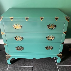 May include: A teal three-drawer chest with brass hardware. The chest has brass corner accents and ornate brass drawer pulls. The legs are curved. The chest is painted in a solid teal color.