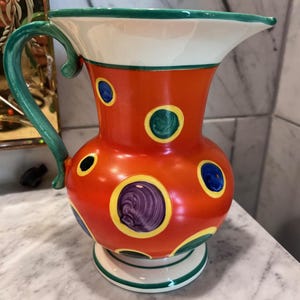 May include: A vibrant, hand-painted ceramic pitcher with an orange body adorned with colorful polka dots. The pitcher features a white rim with a green trim, a green handle, and a white base with green accents. Decorative kitchenware.