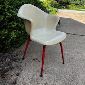 May include: A vintage, off-white molded plastic chair with a curved back and seat. The chair has four bright red metal legs. The chair is designed for comfort and style, suitable for indoor or outdoor use.
