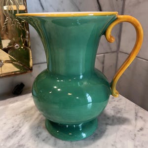 May include: A green ceramic pitcher with a yellow handle and rim. The pitcher has a bulbous body and a flared spout. The handle is curved and attached to the side of the pitcher. The pitcher is sitting on a white surface.