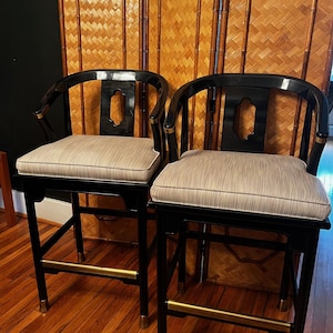 May include: Two black and gold bar stools with a woven back and a beige striped cushion. The stools are in front of a bamboo screen.