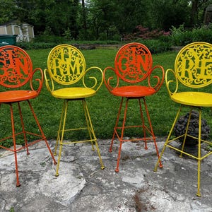 May include: Four colorful metal bar stools. Each stool has a round seat and backrest with a decorative cutout design. The stools are painted in orange and yellow, with the word "GIN" or "RYE" in the backrests.