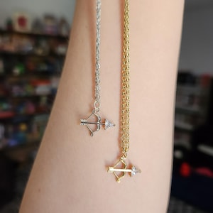 Puede incluir: Dos collares, uno plateado y otro dorado, cada uno con un pequeño colgante de arco y flecha. Las puntas de las flechas son piedras claras y brillantes. Las cadenas son delicadas y cuelgan verticalmente.