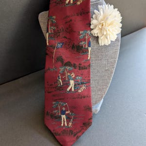 May include: A burgundy tie with a golf course scene featuring golfers, trees, and a flag with the number 15. The tie is displayed on a gray mannequin head with a cream-colored flower.