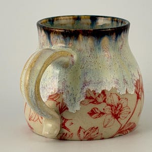 Könnte beinhalten: Handgefertigte Keramik-Tasse mit floralem Muster. Die Tasse hat einen cremefarbenen Grund mit roten Blumenmustern. Der Rand ist dunkelblau, der an den Seiten in eine hellgrüne und violette Glasur übergeht. Der Griff ist cremefarben und goldfarben.