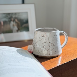 May include: A speckled white ceramic mug with a wide base and a curved handle. The mug features a glossy finish with small brown flecks throughout. A small tea bag tag with the text English Breakfast is visible tucked into the side of the mug.