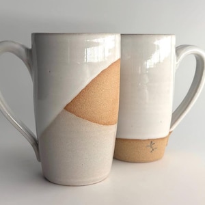 May include: Two tall ceramic mugs with large handles. The mugs are mostly white with a diagonal tan section. The base of the mugs is tan. The mugs are handmade and have a simple, modern design. Perfect for coffee or tea.