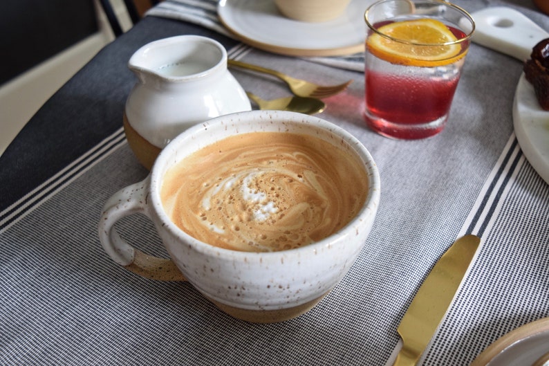 K&ouml;nnte beinhalten: Ein gesprenkelter Keramikbecher gef&uuml;llt mit Kaffee und einem Wirbel aus Sahne an der Oberfl&auml;che. Der Becher hat einen wei&szlig;en oberen Teil und einen beigen Boden. Daneben stehen ein kleines wei&szlig;es Keramik-Milchk&auml;nnchen und ein Glas mit rotem Getr&auml;nk.