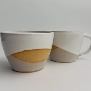 K&ouml;nnte beinhalten: Zwei Keramikbecher mit zweifarbigem Design. Der obere Teil ist mit einer glatten, wei&szlig;en Glasur versehen, w&auml;hrend der untere Bereich eine texturierte, unglasierte, hellbraune Tonoberfl&auml;che mit einer deutlichen, geschwungenen Trennlinie aufweist.