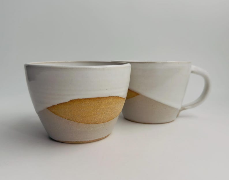 K&ouml;nnte beinhalten: Zwei Keramikbecher mit zweifarbigem Design. Der obere Teil ist mit einer glatten, wei&szlig;en Glasur versehen, w&auml;hrend der untere Bereich eine texturierte, unglasierte, hellbraune Tonoberfl&auml;che mit einer deutlichen, geschwungenen Trennlinie aufweist.