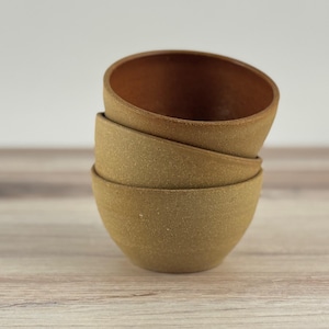 以下が含まれることがあります： 3つの小さなセラミックボウルが積み重ねられています。ボウルは外側が薄いタン色で、内側が濃い茶色です。ボウルは丸みを帯びた形で、互いの上に積み重ねられています。