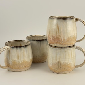 May include: A set of four ceramic mugs with a rounded shape, featuring a cream-colored glaze that transitions into a textured, sandy-brown base and a dark, charcoal-colored rim. The mugs have curved handles and a smooth, glossy finish.