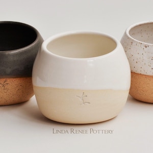 May include: Three rounded ceramic cups with a two-tone design. The left cup is dark gray and tan, the center is white and tan, and the right is white with brown speckles and tan. Each cup features a small stamped mark and the text Linda Renee Pottery.