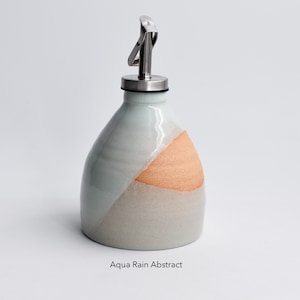 May include: A ceramic oil and vinegar bottle with a stainless steel pour spout. The bottle is glazed in a light blue and beige color with a peach-colored accent.