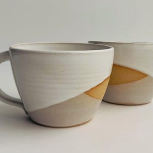 K&ouml;nnte beinhalten: Zwei Keramikbecher mit zweifarbigem Design, bestehend aus einer gl&auml;nzend wei&szlig;en Glasur im oberen Bereich und einer sichtbaren, matten, hellbraunen Tonbasis. Die Becher haben eine breite, abgerundete Schalenform und einen geschwungenen Henkel.