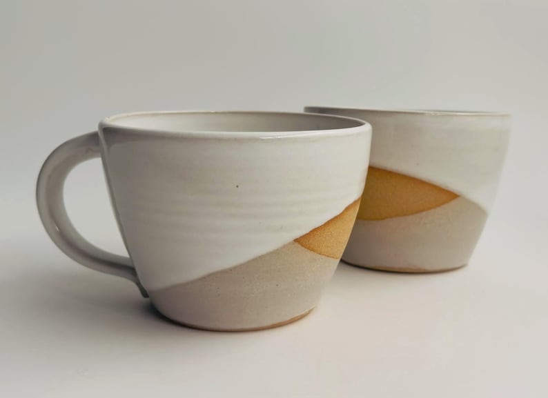 K&ouml;nnte beinhalten: Zwei Keramikbecher mit zweifarbigem Design, bestehend aus einer gl&auml;nzend wei&szlig;en Glasur im oberen Bereich und einer sichtbaren, matten, hellbraunen Tonbasis. Die Becher haben eine breite, abgerundete Schalenform und einen geschwungenen Henkel.