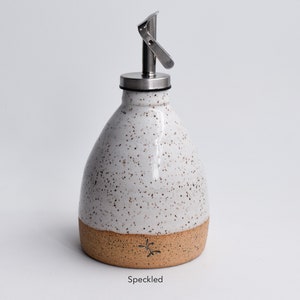 May include: White ceramic oil and vinegar bottle with a speckled glaze and a brown band around the bottom. The bottle has a stainless steel pour spout and lid. "Speckled" is written below the bottle.
