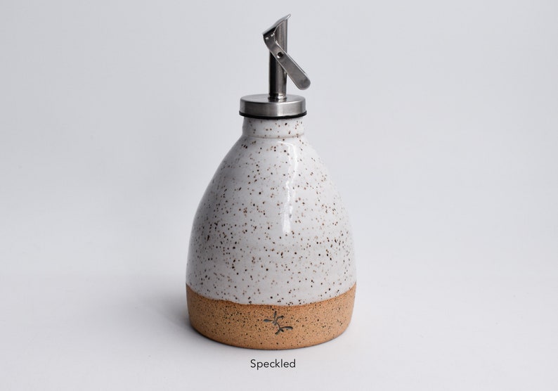 May include: White ceramic oil and vinegar bottle with a speckled glaze and a brown band around the bottom. The bottle has a stainless steel pour spout and lid. "Speckled" is written below the bottle.