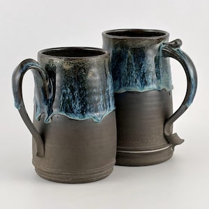 May include: Two handmade ceramic mugs with dark brown bases and handles. The upper portion of each mug features a blue and teal glaze that drips down the sides. The mugs are of different sizes and are set against a white background.