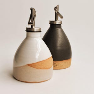 May include: Two ceramic oil dispensers with stainless steel pour spouts. One dispenser features a glossy white upper glaze over a raw, speckled tan base. The second dispenser has a matte black upper glaze over a similar raw, speckled tan base.