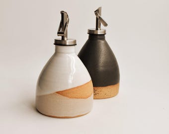 May include: Two ceramic oil dispensers with stainless steel pour spouts. One dispenser features a glossy white upper glaze over a raw, speckled tan base. The second dispenser has a matte black upper glaze over a similar raw, speckled tan base.