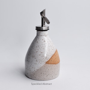 May include: White ceramic oil and vinegar bottle with a speckled glaze and a brown accent. The bottle has a stainless steel pour spout with a lever.
