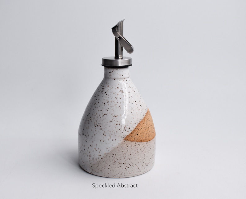 May include: White ceramic oil and vinegar bottle with a speckled glaze and a brown accent. The bottle has a stainless steel pour spout with a lever.