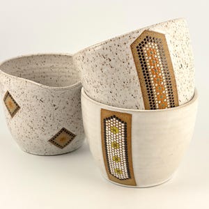 May include: Two ceramic bowls with a speckled off-white finish. One bowl features a vertical rectangular panel with orange and teal dot patterns. The other bowl has a speckled surface with two small diamond-shaped accents featuring brown and yellow dots.