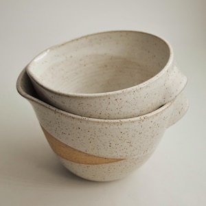 May include: Two stacked ceramic bowls with a speckled off-white glaze and a smooth, unglazed tan band around the lower exterior. Each bowl features a wide, rounded opening and a small, protruding handle on the side for a secure grip.