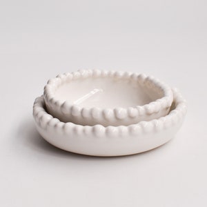May include: Two white ceramic bowls with a beaded rim. The bowls are nested together.