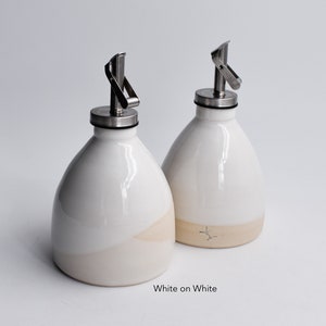 May include: Two white ceramic oil and vinegar bottles with stainless steel pour spouts. The bottles have a white glaze on the top half and a cream glaze on the bottom half. The text "White on White" is printed below the bottles.